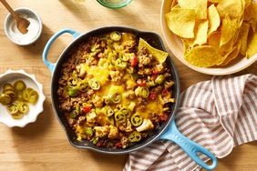 Cheesy ground beef & cauliflower casserole recipe with one tortilla chip scooping casserole out, a bowl of more chips is on the side of the casserole skillet