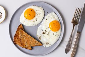 Pristine Sunny-Side Up Eggs sprinkled with salt and pepper and served with whole wheat toast on a plate with fork and knife